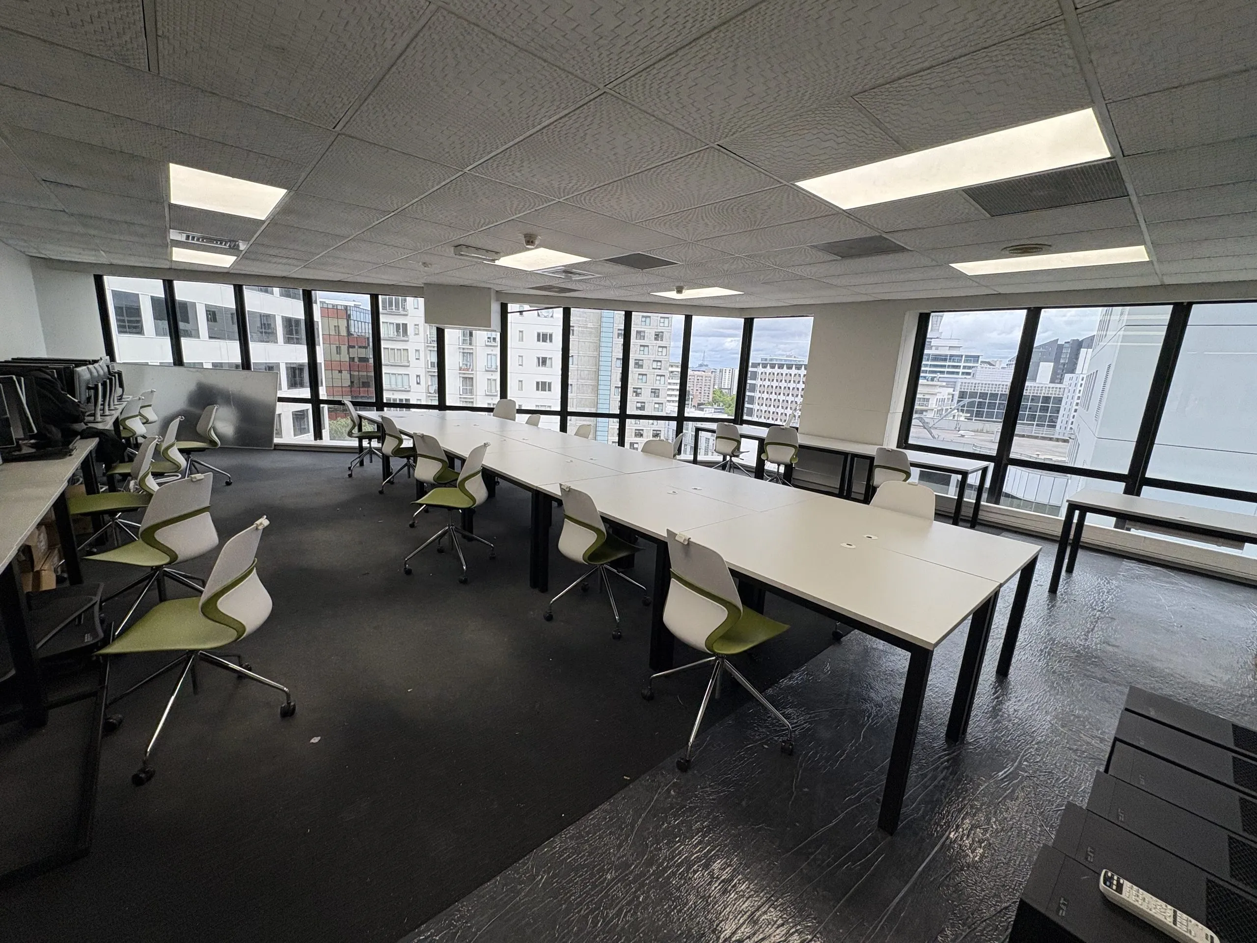 Wide room with installed white tables, green and white chairs and a wall of windows.