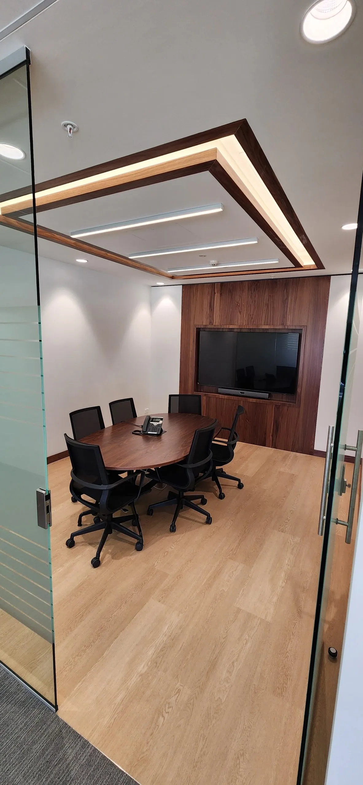 Meeting room with a timber table, black chairs, a wall-mounted screen and a lit ceiling tray.
